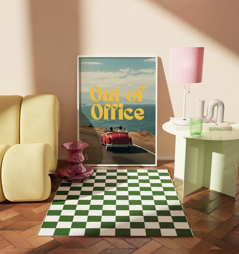 May include: A framed print with a retro aesthetic featuring a red car driving on a winding road with the text "Out of Office" in yellow letters. The print is hanging on a wall above a green and white checkered rug.