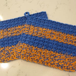 May include: Two handmade, blue and orange striped potholders. The potholders are made of a thick, textured yarn and have a loop for hanging. The color scheme is a vibrant combination of blue and orange.