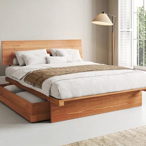 May include: A platform bed frame made of light brown wood with a horizontal slatted headboard and a built-in storage drawer at the base. The bed is dressed with white linens, white pillows, and a brown textured throw blanket draped across the foot.