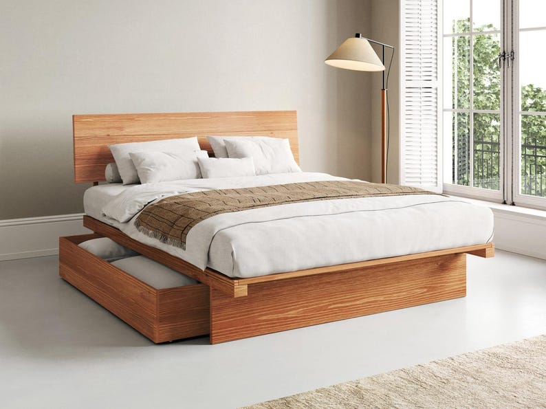 May include: A platform bed frame made of light brown wood with a horizontal slatted headboard and a built-in storage drawer at the base. The bed is dressed with white linens, white pillows, and a brown textured throw blanket draped across the foot.