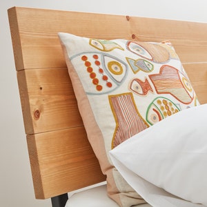 May include: A square throw cushion featuring an off-white fabric base with colourful embroidered fish motifs in shades of orange, green, and blue, resting against a horizontal light-toned wooden headboard next to a plain white pillow.