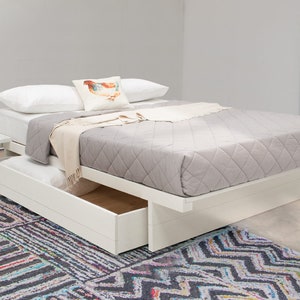 May include: A white platform bed frame with a built-in side shelf and a large pull-out storage drawer underneath. The bed is topped with a gray quilted mattress cover, two white pillows, and a small decorative pillow featuring a chicken illustration.