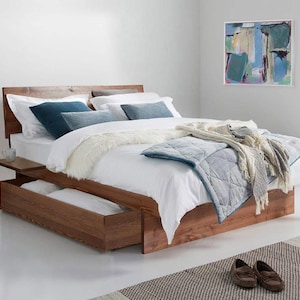 May include: A wooden platform bed frame with a headboard and integrated side shelf. The bed features a large pull-out storage drawer at the base, dressed with white bedding, a blue quilted coverlet, and several blue and white decorative pillows.