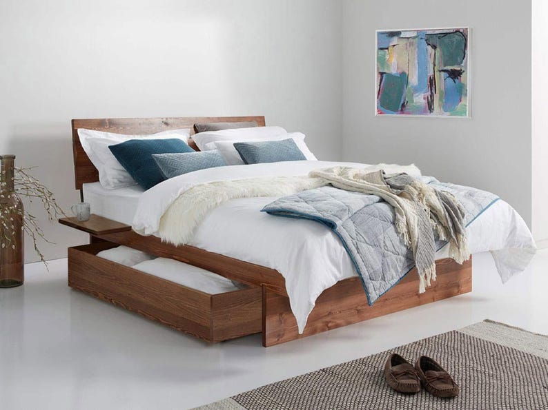 May include: A wooden platform bed frame with a headboard and integrated side shelf. The bed features a large pull-out storage drawer at the base, dressed with white bedding, a blue quilted coverlet, and several blue and white decorative pillows.