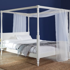 May include: White wooden four-poster bed frame with a white canopy and white bedding. The bed is on a light brown rug in front of a dark blue wall.