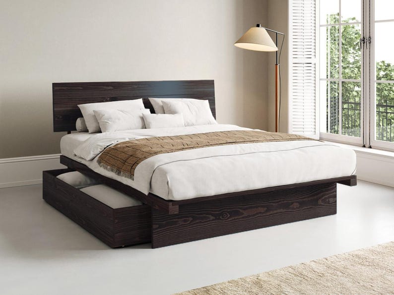 May include: A dark brown wooden platform bed frame with a rectangular headboard and a low-profile base. The bed features a pull-out storage drawer on the side, topped with a white mattress, white pillows, and a brown textured throw blanket.