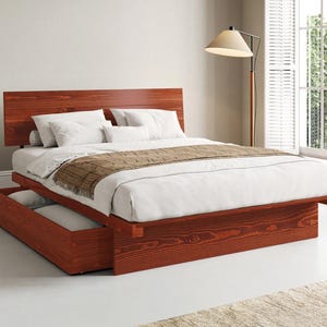 May include: A platform bed frame made of reddish-brown wood with a tall headboard and a built-in storage drawer at the base. The bed is dressed with white linens, white pillows, and a brown textured runner draped across the foot of the mattress.