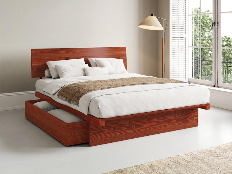 May include: A platform bed frame made of reddish-brown wood with a tall headboard and a built-in storage drawer at the base. The bed is dressed with white linens, white pillows, and a brown textured runner draped across the foot of the mattress.