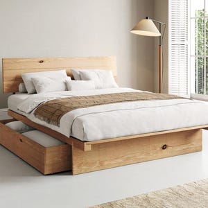 May include: A light-toned wooden platform bed frame featuring a horizontal plank headboard and a built-in storage drawer at the base. The bed is dressed with white linens, two white pillows, and a brown plaid runner draped across the foot.