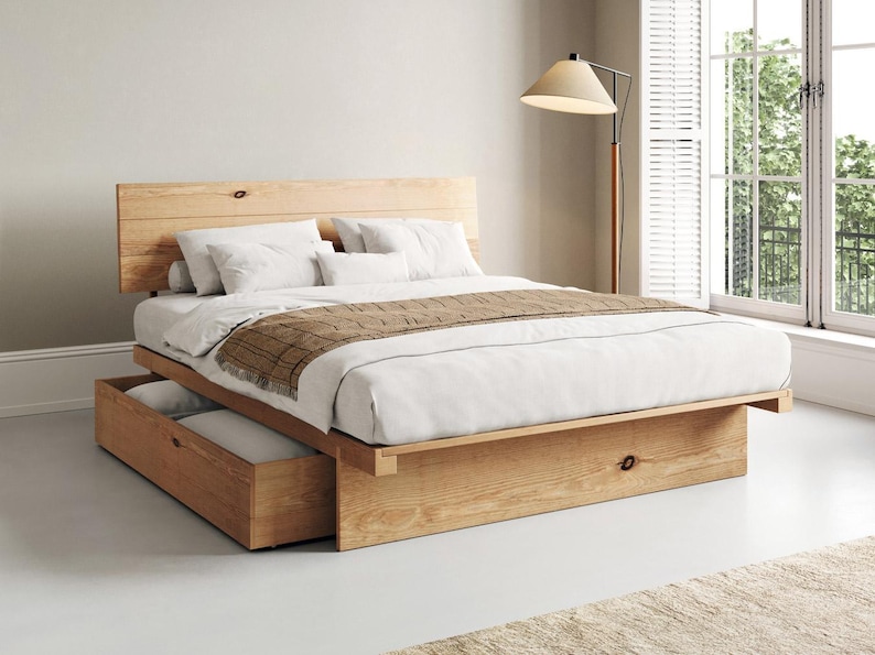 May include: A light-toned wooden platform bed frame featuring a horizontal plank headboard and a built-in storage drawer at the base. The bed is dressed with white linens, two white pillows, and a brown plaid runner draped across the foot.
