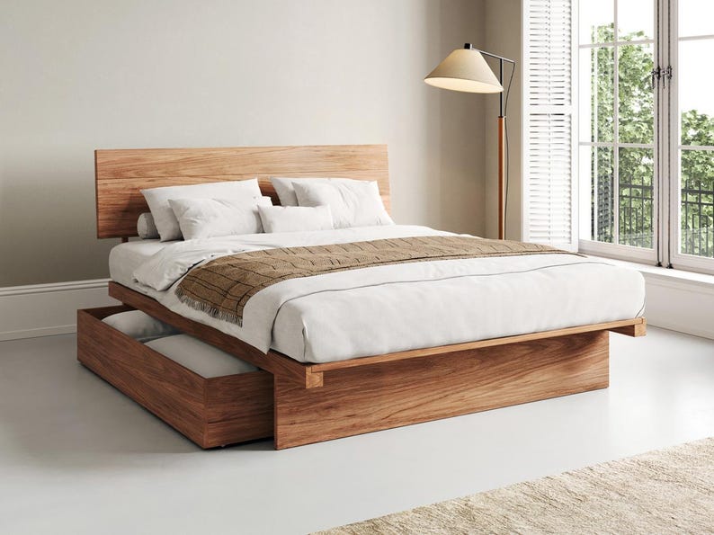 May include: A platform bed frame crafted from light brown wood with a prominent headboard and a built-in storage drawer at the base. The bed is dressed with white linens, two white pillows, a small bolster pillow, and a brown textured runner.