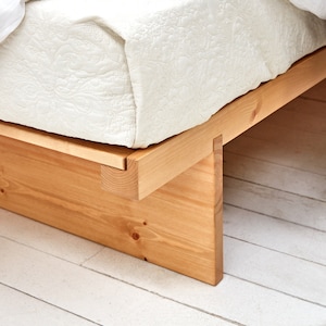 May include: A low-profile wooden bed frame constructed from light-coloured natural wood planks with visible grain and knots. The frame features a simple joinery design supporting a white quilted mattress and a patterned throw blanket on the side.