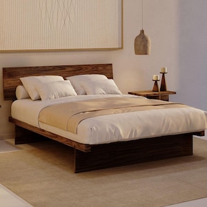 May include: A low-profile platform bed frame crafted from dark brown wood with a prominent grain pattern. The bed features a rectangular headboard and a solid base, dressed with white pillows and a white duvet cover with a light tan runner.