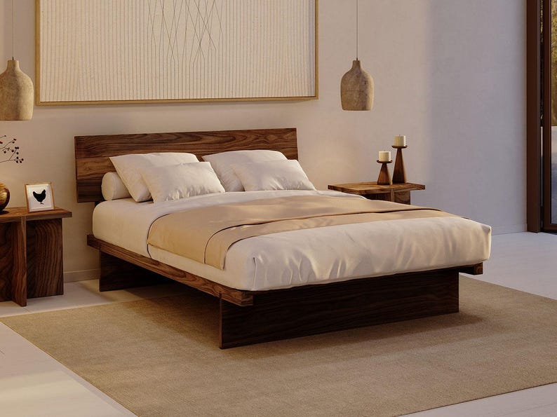 May include: A low-profile platform bed frame crafted from dark brown wood with a prominent grain pattern. The bed features a rectangular headboard and a solid base, dressed with white pillows and a white duvet cover with a light tan runner.