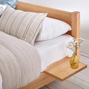 May include: A wooden bed frame with a white duvet and pillows. The bed has a small wooden shelf attached to the side, with a vase of white flowers on it.