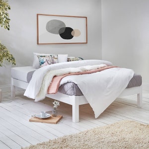 Op de afbeelding: Wit platform bedframe met een wit dekbed en een roze deken. Het bed is opgemaakt met witte lakens en heeft twee kussens met bloemenpatronen.