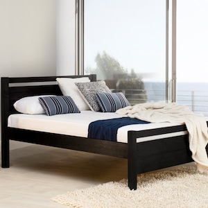 May include: A black wooden bed frame with a slatted headboard and footboard. The bed is dressed with a white mattress, two white pillows, two blue and white striped rectangular pillows, one grey patterned pillow, and a cream-colored knit throw.