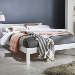 May include: A white wooden bed frame with a headboard, featuring a white mattress topped with a dark gray quilt, a light pink quilted blanket, and a gray and white striped throw blanket with tassels. Two pillows are placed against the headboard.
