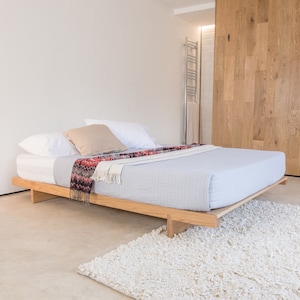 Op de afbeelding: Een licht houten platformbedframe met een lichtblauwe bedsprei en een gestreepte deken. Het bed is opgemaakt met twee witte kussens en een beige kussen.