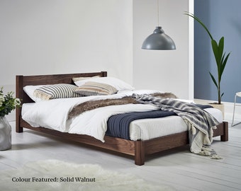Op de afbeelding: Een bedframe van massief walnoothout met een laag profiel en een rechthoekig hoofdeinde. Het bed is opgemaakt met wit beddengoed, een donkerblauwe doorgestikte sprei, een imitatiebont loper en twee gestreepte sierkussens. Tekst: Colour Featured: Solid Walnut.