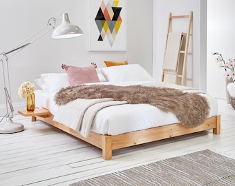 May include: A low-profile light wood platform bed frame with an integrated side table, dressed in white bedding, a beige throw blanket, and a textured brown faux fur runner, accented by a pink pom-pom pillow and a mustard yellow velvet pillow.