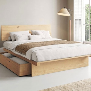 May include: A light-coloured wooden platform bed frame with a horizontal plank headboard and a built-in storage drawer underneath. The bed is dressed with white linens, white pillows, and a brown textured runner draped across the foot of the bed.