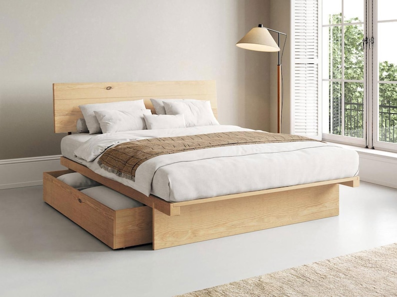 May include: A light-coloured wooden platform bed frame with a horizontal plank headboard and a built-in storage drawer underneath. The bed is dressed with white linens, white pillows, and a brown textured runner draped across the foot of the bed.