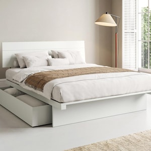 May include: A white platform bed frame with a horizontal panelled headboard and a large pull-out storage drawer underneath. The bed is dressed with white bedding, several white pillows, and a brown textured runner draped across the foot.