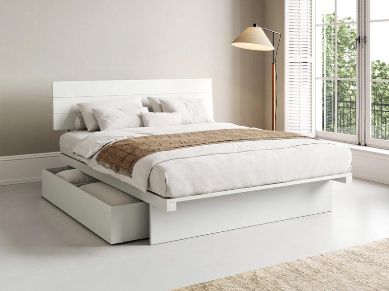 May include: A white platform bed frame with a horizontal panelled headboard and a large pull-out storage drawer underneath. The bed is dressed with white bedding, several white pillows, and a brown textured runner draped across the foot.