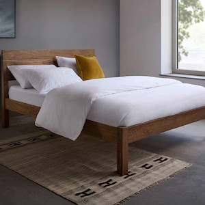May include: A wooden bed frame with a natural brown finish and a tall headboard. The bed is dressed with white bedding, including a duvet and two pillows, and features a single mustard yellow square accent pillow placed against the headboard.