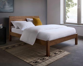 May include: A wooden bed frame with a natural brown finish and a tall headboard. The bed is dressed with white bedding, including a duvet and two pillows, and features a single mustard yellow square accent pillow placed against the headboard.