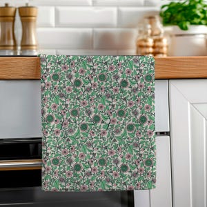 May include: A patterned kitchen towel featuring a floral design. The pattern includes pink flowers, green leaves, and dark stems against a light backdrop. The towel is displayed in a kitchen setting, hanging on a wooden surface.