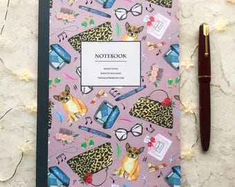 May include: A notebook with a pink cover featuring illustrations of a chihuahua, sunglasses, purses, and perfume bottles. The notebook has a dark green spine and the text "NOTEBOOK" and "MADE IN ENGLAND". A burgundy fountain pen is next to the notebook.