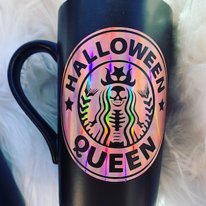 以下が含まれることがあります： 黒いセラミック製のマグカップに、王冠をかぶった骸骨と「Halloween Queen」の文字が書かれたホログラフィックステッカーが貼られています。