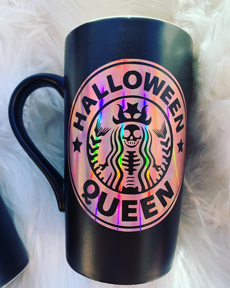 以下が含まれることがあります： 黒いセラミック製のマグカップに、王冠をかぶった骸骨と「Halloween Queen」の文字が書かれたホログラフィックステッカーが貼られています。