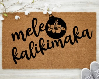 May include: A brown coir doormat with the text "mele kalikimaka" in black cursive lettering. A black silhouette of a Christmas ornament with a hibiscus flower inside is centered above the text.