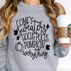 May include: A light gray sweatshirt with the words "Comfy Sweaters Hocus Pocus Pumpkin Everything" in black script. The design includes leaf and pumpkin graphics. A white coffee cup with a brown sleeve is held in the hand.