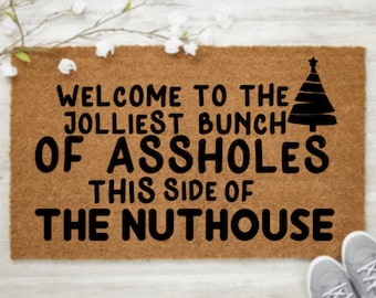 May include: A rectangular doormat with the text "WELCOME TO THE JOLLIEST BUNCH OF ASSHOLES THIS SIDE OF THE NUTHOUSE" in black. A black Christmas tree graphic is on the right. A pair of gray sneakers is at the bottom right.