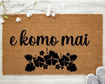 May include: A brown coir doormat with the text "e komo mai" in black cursive lettering. The text is surrounded by a row of black hibiscus flowers and leaves.