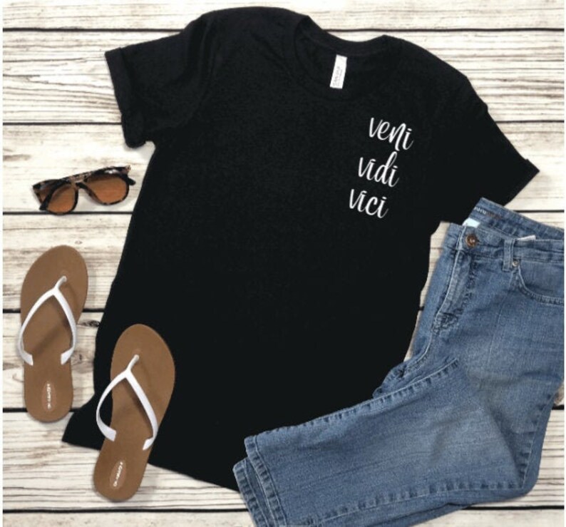 Op de afbeelding: Zwart T-shirt met de woorden "veni vidi vici" in witte letters. Het shirt wordt gecombineerd met een blauwe spijkerbroek, bruine slippers met witte bandjes en een zonnebril. De items zijn gerangschikt op een licht houten oppervlak.