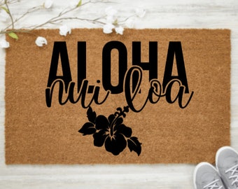 May include: A rectangular brown coir doormat with the words "ALOHA nui loa" in black script. A black hibiscus flower design is below the text. The mat is decorated with white flowers and a pair of gray sneakers.