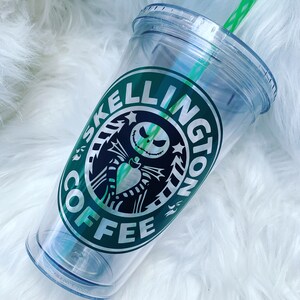 May include: Clear plastic tumbler with a green straw and lid. The tumbler features a green and black logo that says "Skelington Coffee" with a Jack Skellington design. The tumbler is on a white, fluffy surface.