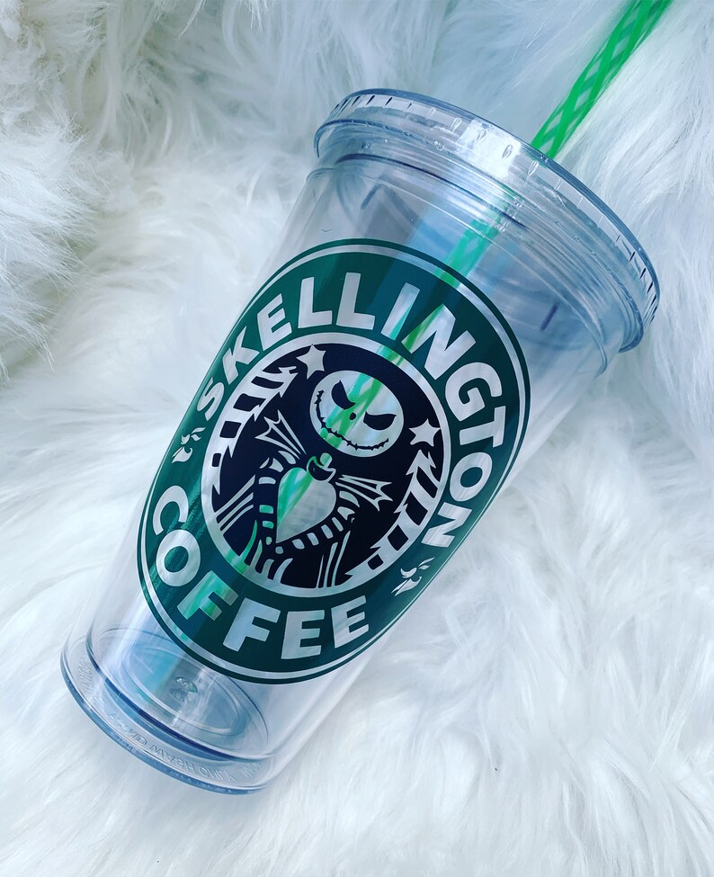 May include: Clear plastic tumbler with a green straw and lid. The tumbler features a green and black logo that says "Skelington Coffee" with a Jack Skellington design. The tumbler is on a white, fluffy surface.