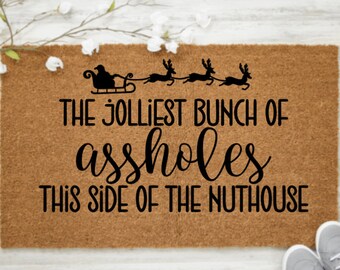 May include: A rectangular doormat made of natural coir fiber. The mat features a black silhouette of Santa's sleigh and reindeer, with the text "THE JOLLIEST BUNCH OF assholes THIS SIDE OF THE NUTHOUSE" in a playful font.