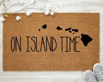May include: A rectangular doormat made of brown coir fiber. The mat features a black silhouette of the Hawaiian Islands and the phrase "ON ISLAND TIME" in a bold, black font. A pair of gray sneakers is visible in the bottom right corner.