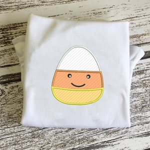May include: White t-shirt with an embroidered candy corn design. The candy corn has a yellow, orange, and white striped pattern and a smiling face.