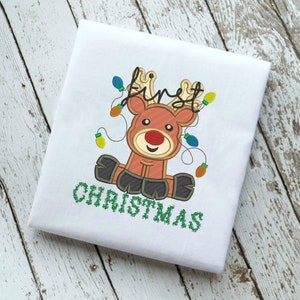 May include: A white onesie with an embroidered design of a red reindeer with brown antlers and a red nose. The reindeer is surrounded by colorful lights and the text "first christmas" is embroidered below the reindeer.