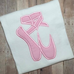 May include: Pink ballet shoes with a pink ribbon embroidered on a white t-shirt.