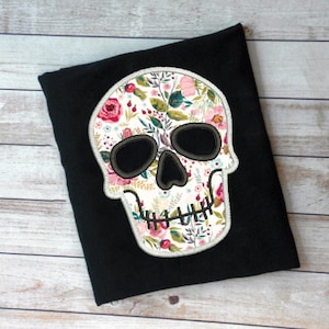 May include: A black t-shirt featuring a white floral skull design. The skull is outlined in grey and has a floral pattern inside.