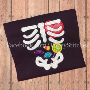 May include: A black t-shirt with a white embroidered skeleton design. The skeleton's heart is filled with a red candy heart. The skeleton's stomach is filled with candy, including a lollipop, candy corn, and wrapped candies.